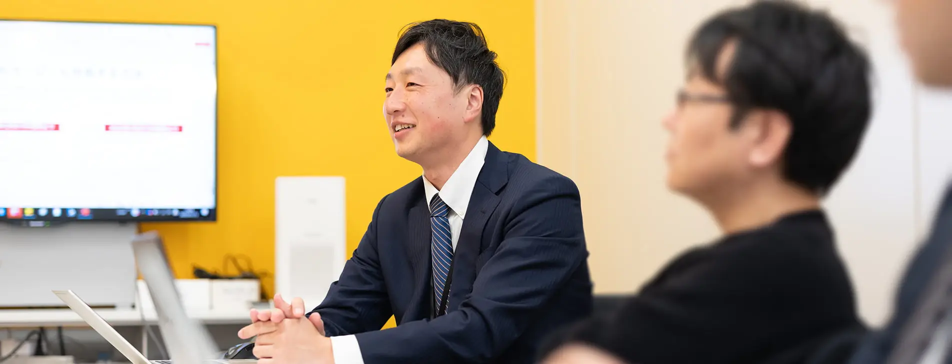 黄色い壁を背景に、会議室でノートパソコンを前に笑顔で話すスーツ姿の男性ビジネスパーソン。背景には大型ディスプレイが設置され、手前にはぼやけた形で別の参加者が映っており、和やかな雰囲気のミーティングの一場面が捉えられている。